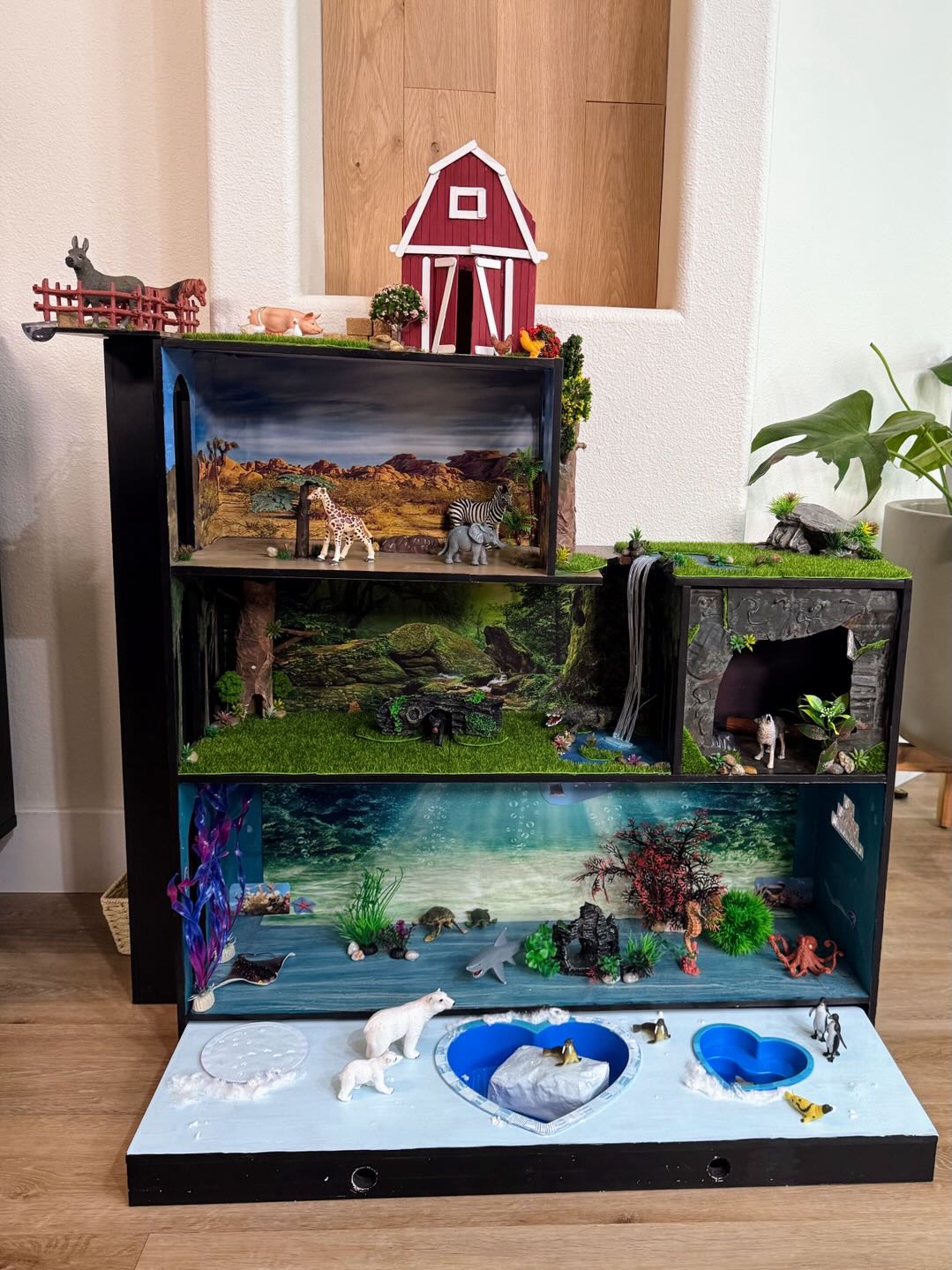 A four-level handmade play habitat: a red barn on top, a desert savanna with giraffe and zebra, a jungle with a waterfall and cave, and an underwater ocean scene with an arctic shelf below