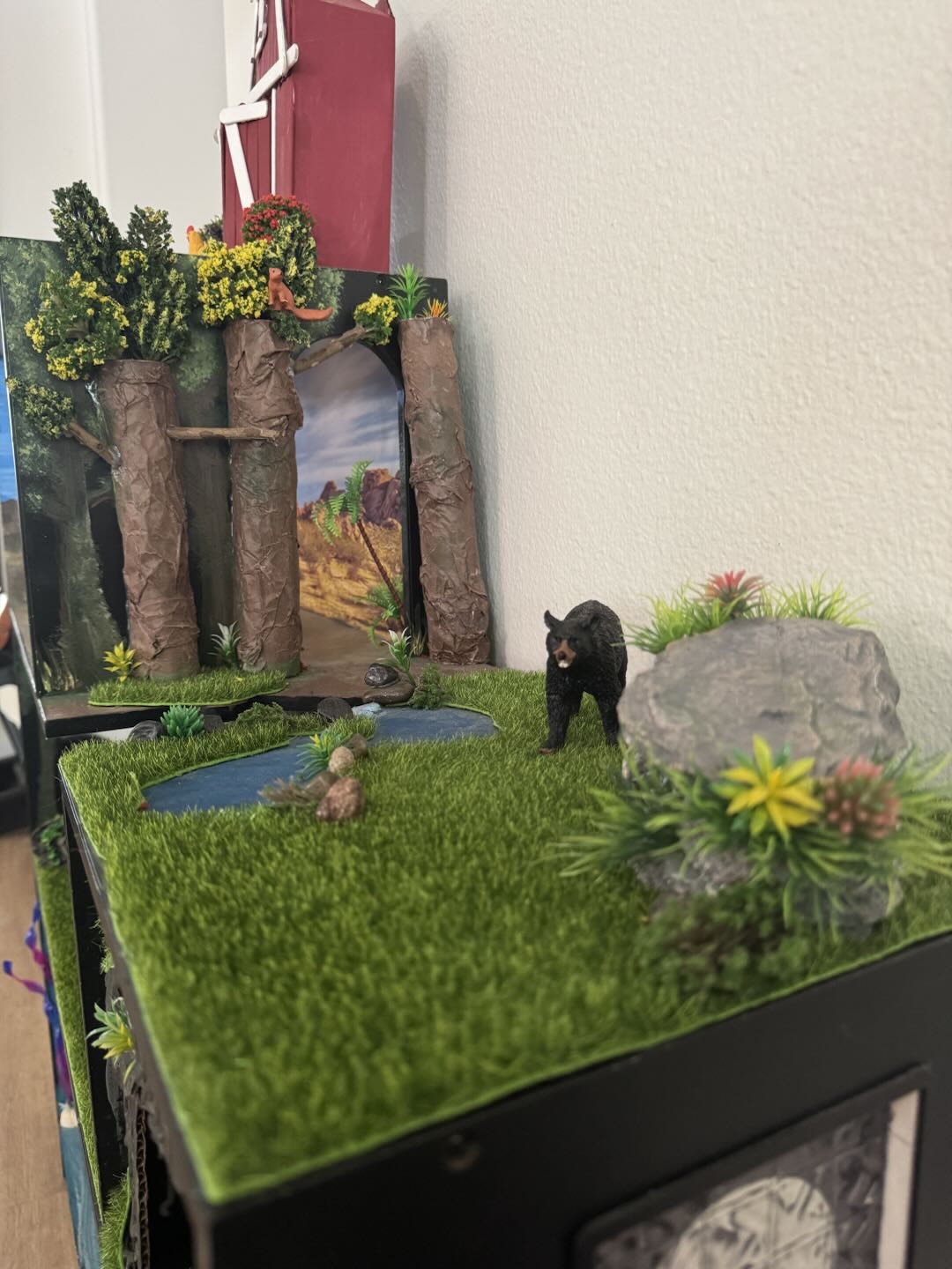 A grassy meadow level with a black bear, a winding blue pond, and a flowering boulder
